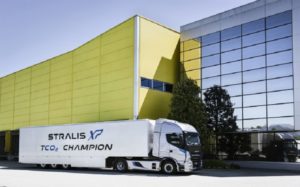 Bílý kamion s návěsem Stralis Champion před moderní budovou ze skla